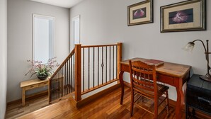 Une pièce avec un bureau en bois, une chaise et une petite table sur laquelle se trouve une lampe. On y trouve également un escalier avec une rampe en bois et une plante en pot posée sur le sol.
