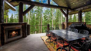 Terraza de madera con chimenea, mesa y sillas al aire libre, con vistas a un bosque.