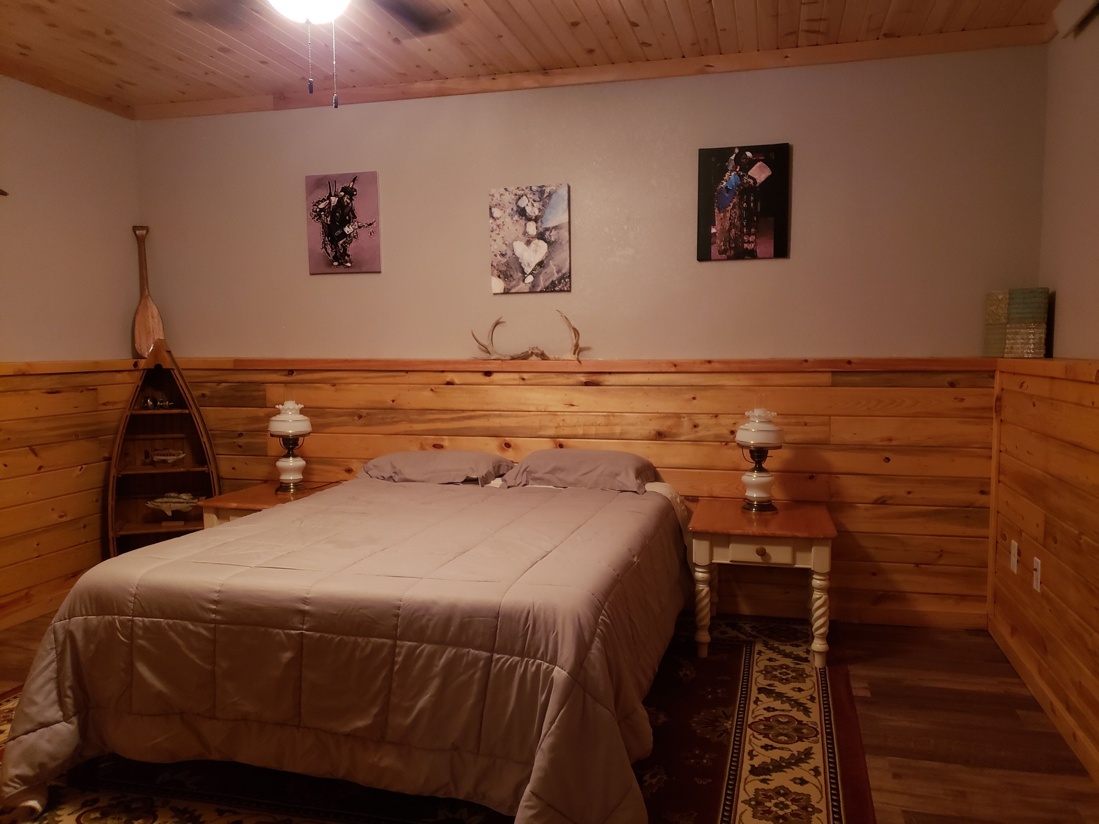 A cozy room with a wooden headboard, a bed with a beige comforter, two bedside tables with lamps, and three wall-mounted artworks.