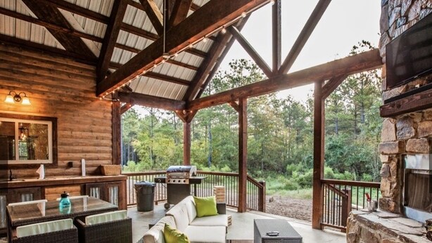 A covered outdoor patio with wicker furniture, a stone fireplace, and a view of the surrounding forest. - Romantic Cabin in Broken Bow! Secluded on 4+ acres, 1BR 2Bath, Hot Tub, FirePit (Broken Bow)
