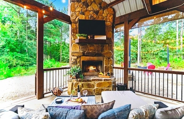 A stone fireplace with a TV above it, surrounded by outdoor seating and a view of a forest.