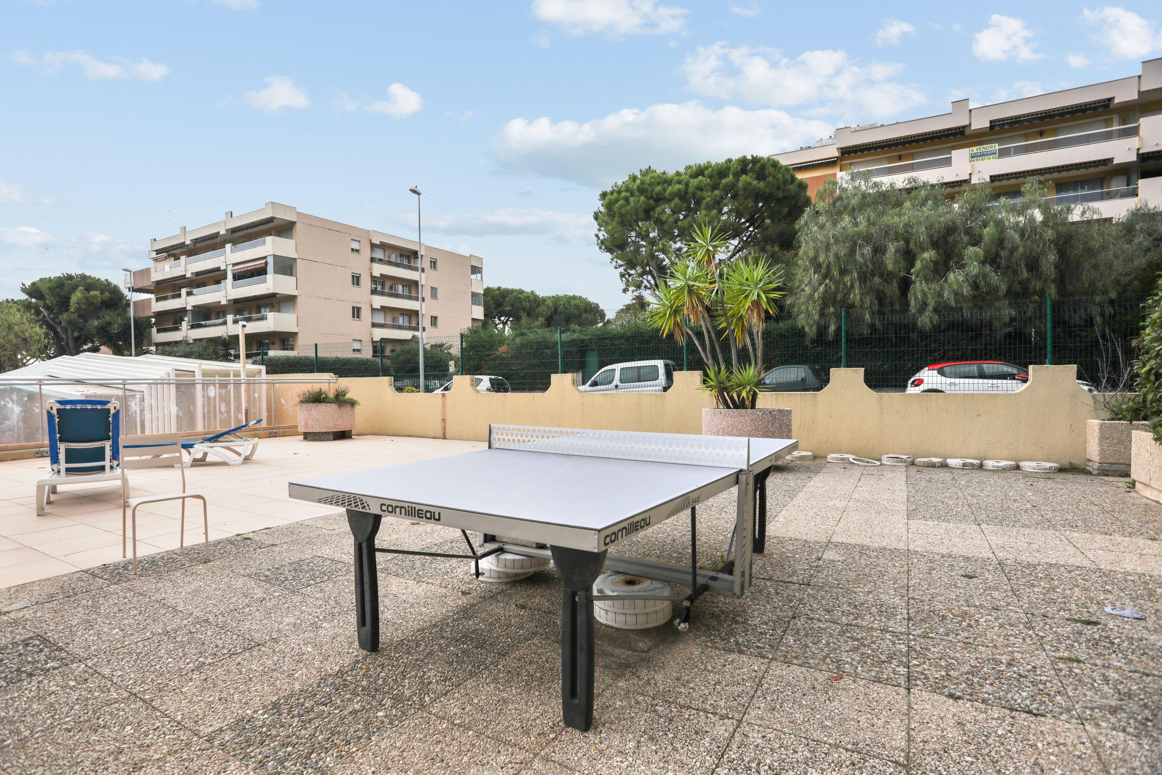 A ping pong table in an outdoor area with a paved surface, surrounded by potted plants and a fence.