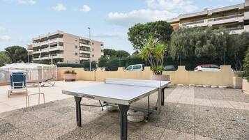 A ping pong table in an outdoor area with a paved surface, surrounded by potted plants and a fence.