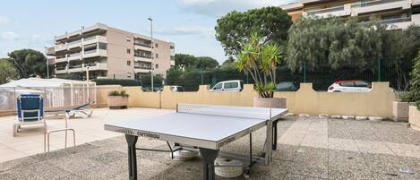A ping pong table in an outdoor area with a paved surface, surrounded by potted plants and a fence.