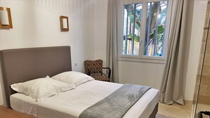 A neatly made bed with white linens and a grey throw, a wooden nightstand, a chair with a patterned cushion, and a window with curtains. - Chez Simone ( 6 pers.) (Ensuès-la-Redonne)