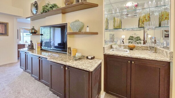 A kitchen with dark wooden cabinets, a granite countertop, and a built-in TV. There are shelves above the counter holding decorative items and glassware. - Corner 2-bed / 2-bath Unit #926, in the Stoneybrook Golf and Country Club (Sarasota)