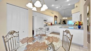 A modern kitchen with white cabinets, a central island, and a dining area with a glass table and metal chairs. - Corner 2-bed / 2-bath Unit #926, in the Stoneybrook Golf and Country Club (Sarasota)
