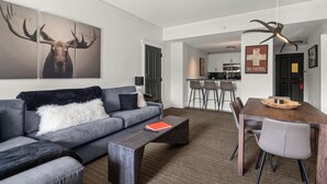 A modern living room with a grey sofa, a coffee table, and a dining area with a wooden table and chairs. There is a kitchen in the background with white cabinets and a black door.