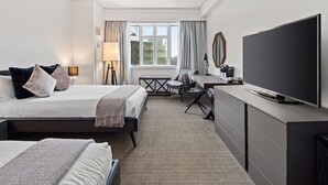 A hotel room with two beds, a desk, a TV, and a view of buildings through the window.