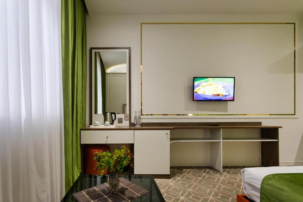 A hotel room with a bed, a desk with a mirror, a television mounted on the wall, and a small table with a plant.