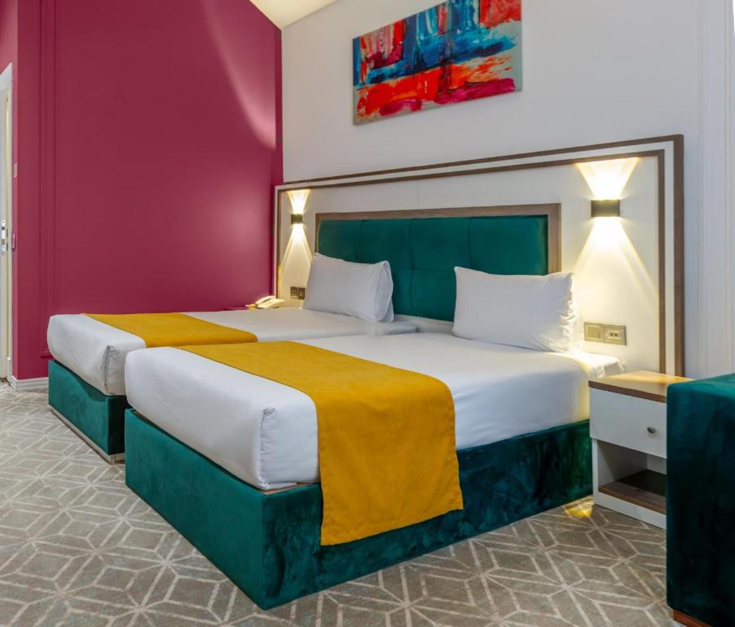 A hotel room with a double bed, two bedside tables, a nightstand, a painting on the wall, and a patterned floor.