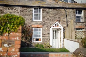 A stone house with a white door and windows, surrounded by a brick wall and greenery.