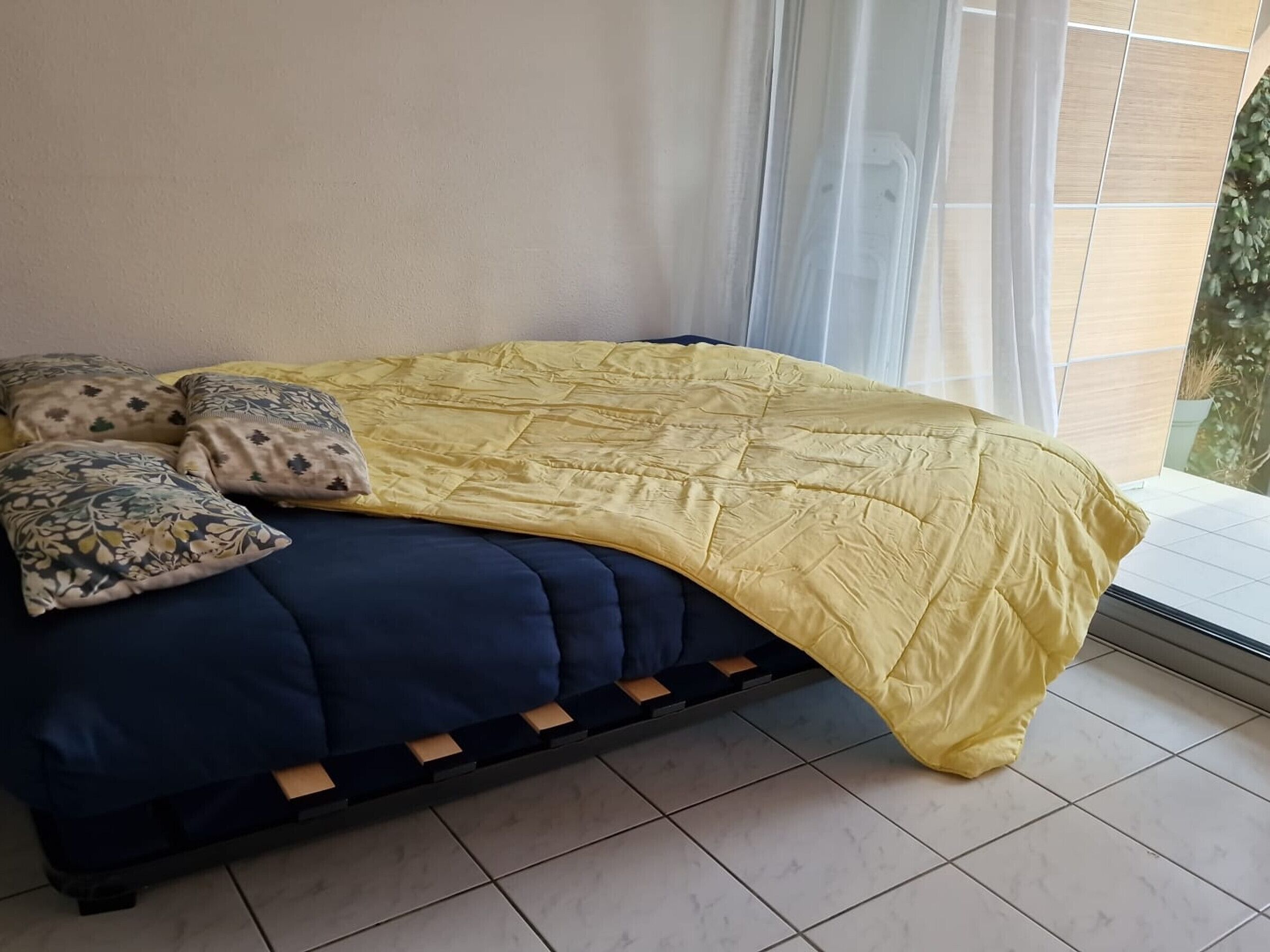 Ein Bett mit blauer Matratze und einer gelben Decke, zwei gemusterte Kissen und ein durchsichtiger Vorhang dahinter.