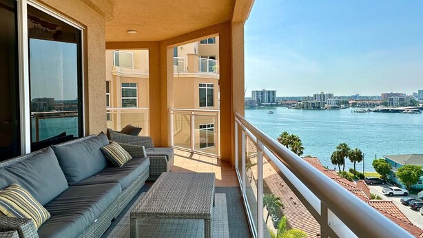 A balcony with a sofa and a small table, overlooking a waterfront with buildings and boats.