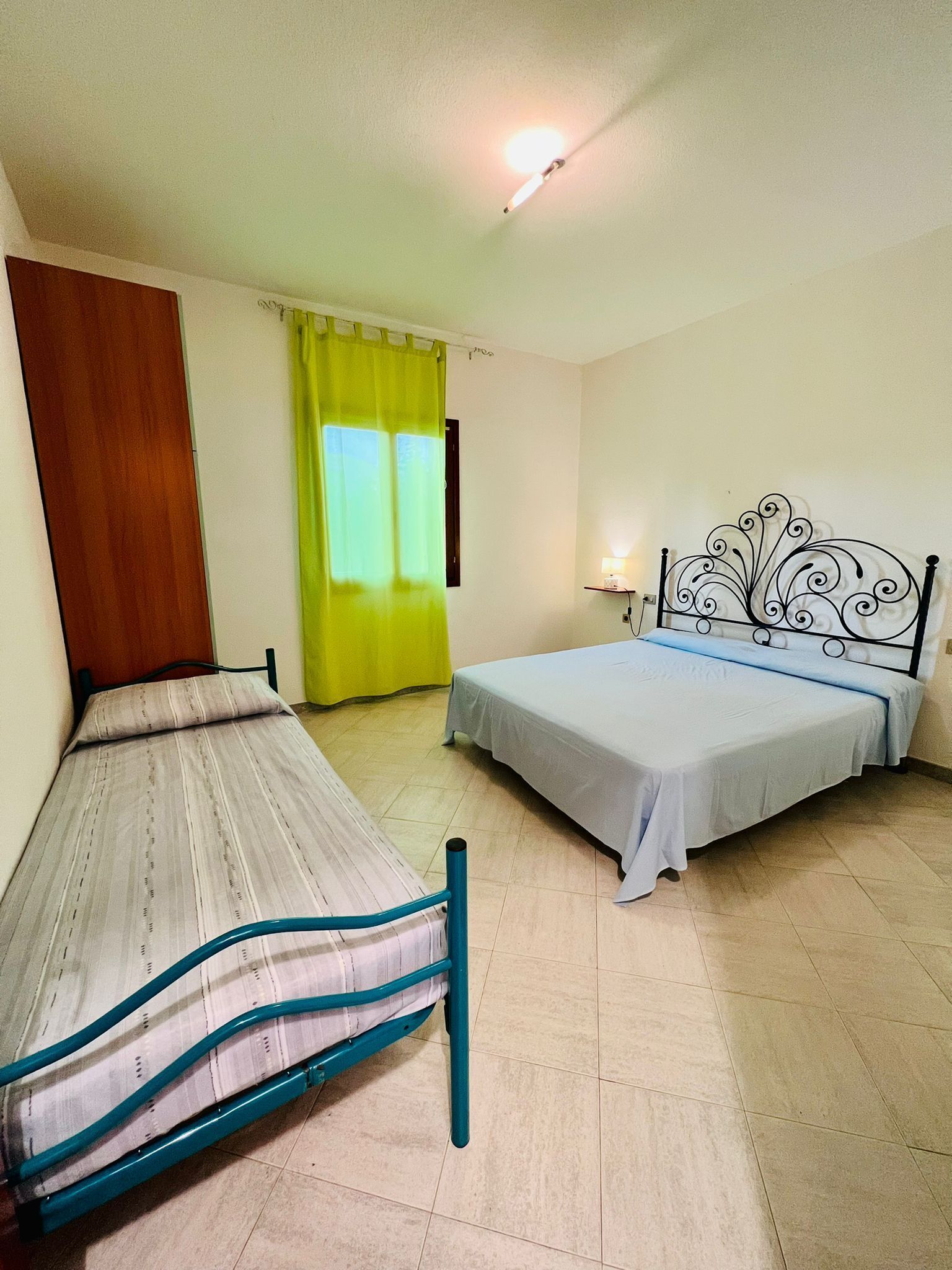 Une petite pièce simple, avec deux lits simples, une porte en bois, un rideau jaune vif et un luminaire au plafond.