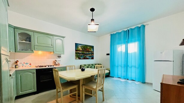 A compact kitchen with a dining area, featuring light green cabinets, a tiled floor, a painting on the wall, and a blue curtain. - Shardana 2 (San Teodoro)