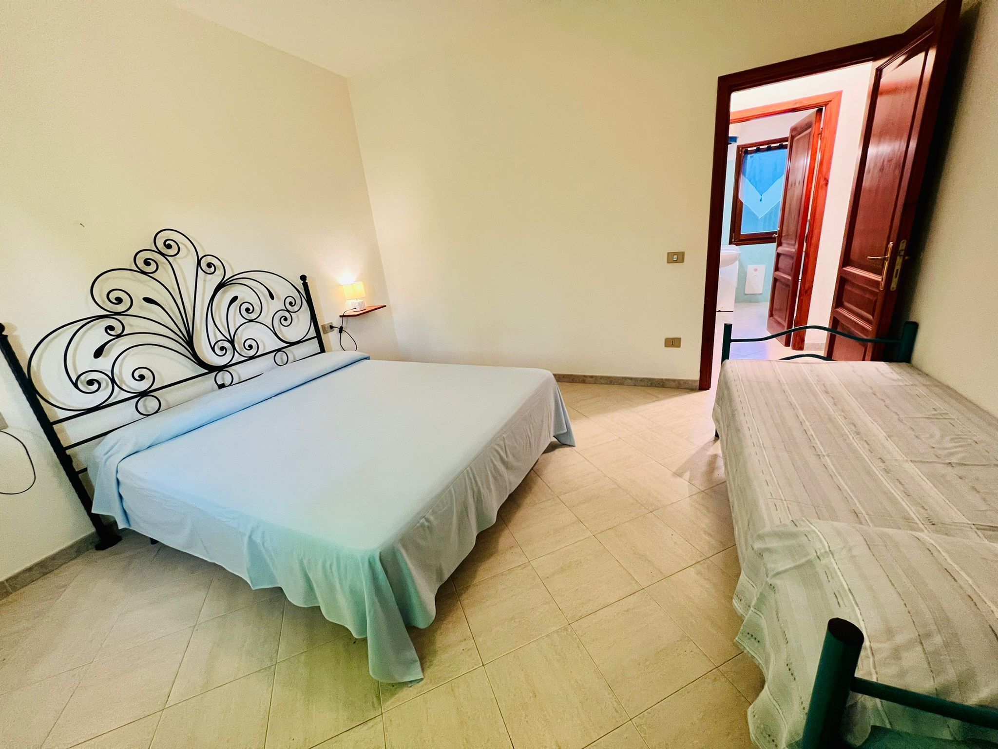 A bedroom with two beds, a nightstand with a lamp, and a wooden door leading to another room.
