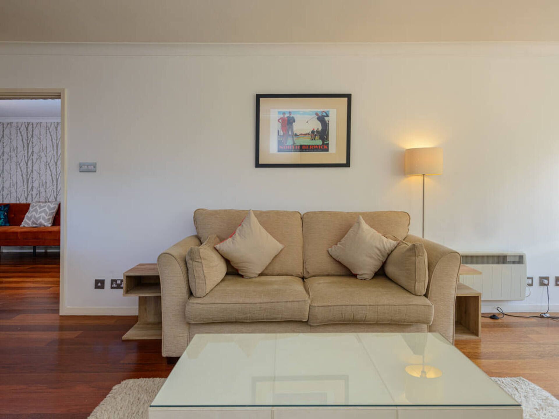 A living room with a beige sofa, a glass coffee table, a framed picture on the wall, and a floor lamp.