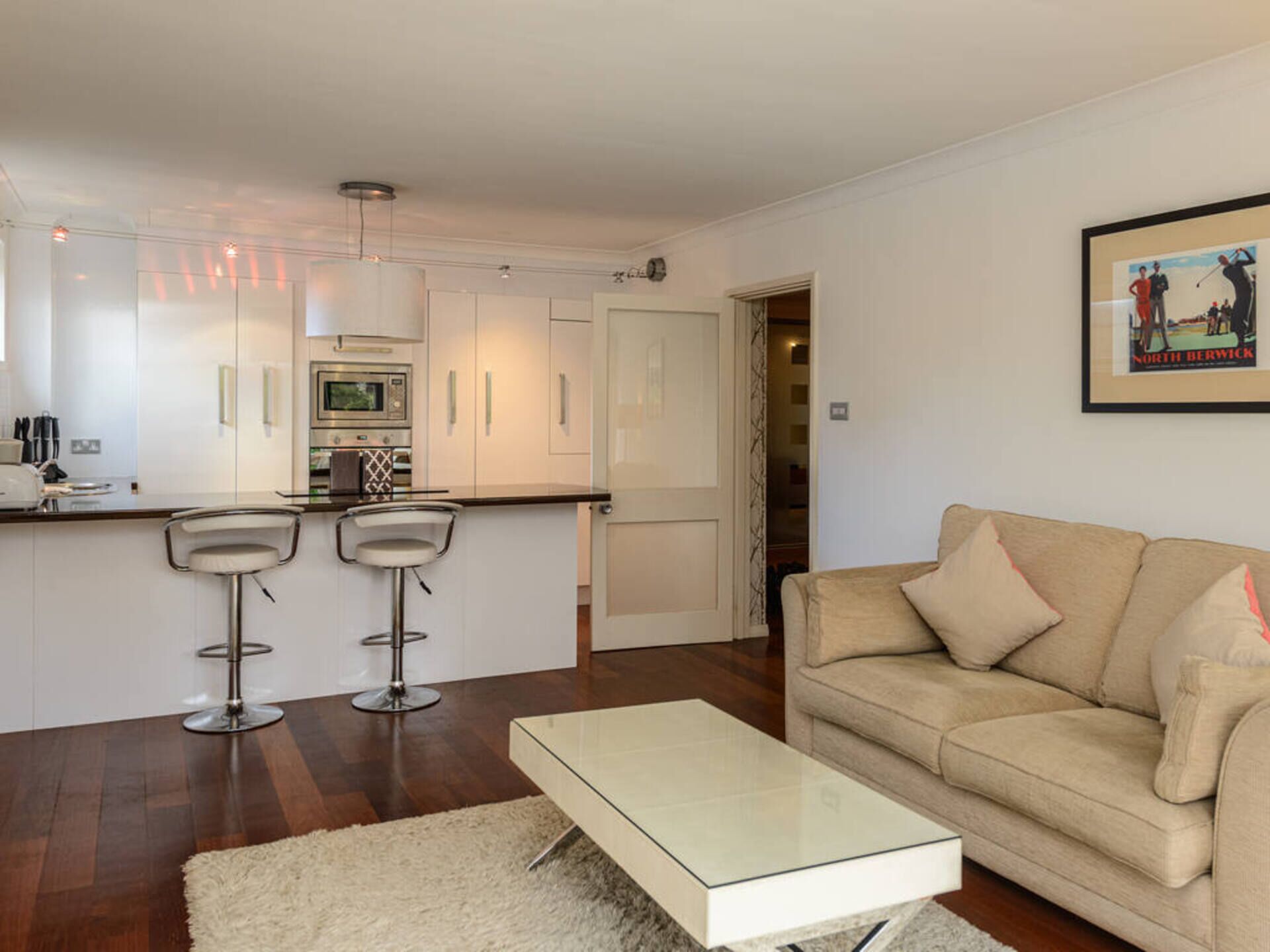 Eine moderne Küche mit weißen Schränken, einer zentralen Kochinsel mit Barhocker, einem Essbereich mit beiger Couch und einem gerahmten Bild an der Wand.