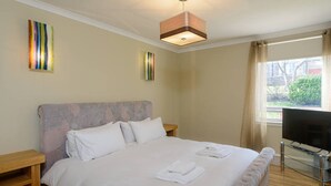 A neatly made bed with white linens and towels, a wooden nightstand, a glass TV stand, and a ceiling light fixture. - Corrin - Chic 1 Bedroom Apartment (North Berwick)