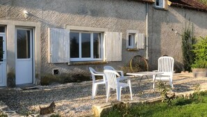 Una casa rustica con un patio ghiaioso, sedie in plastica bianca e un tavolo di legno.
