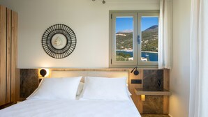 Uma cama bem arrumada com lençóis brancos, um candeeiro circular fixado na parede e uma janela com vista para uma paisagem montanhosa.
