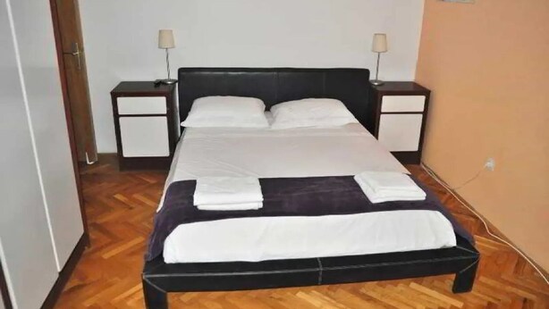 A single bed with a black headboard and white bedding, flanked by two bedside tables with lamps, in a room with wooden flooring. - Park Apartment (Split)