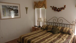 Un dormitorio con una cama, una mesita de noche, una lámpara, un cuadro enmarcado, una ventana con persianas y un adorno en la pared.