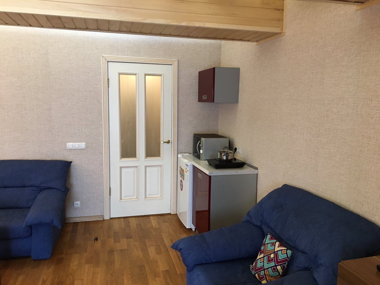 A room with two blue sofas, a wooden ceiling, a white door with frosted glass panels, a small kitchenette with a microwave and a pot, and a cabinet above it.