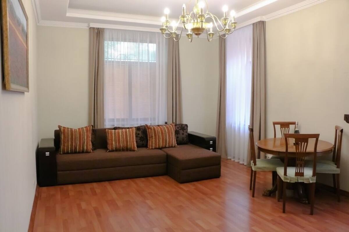A living room with a sectional sofa, a dining table with chairs, a chandelier, and sheer curtains.