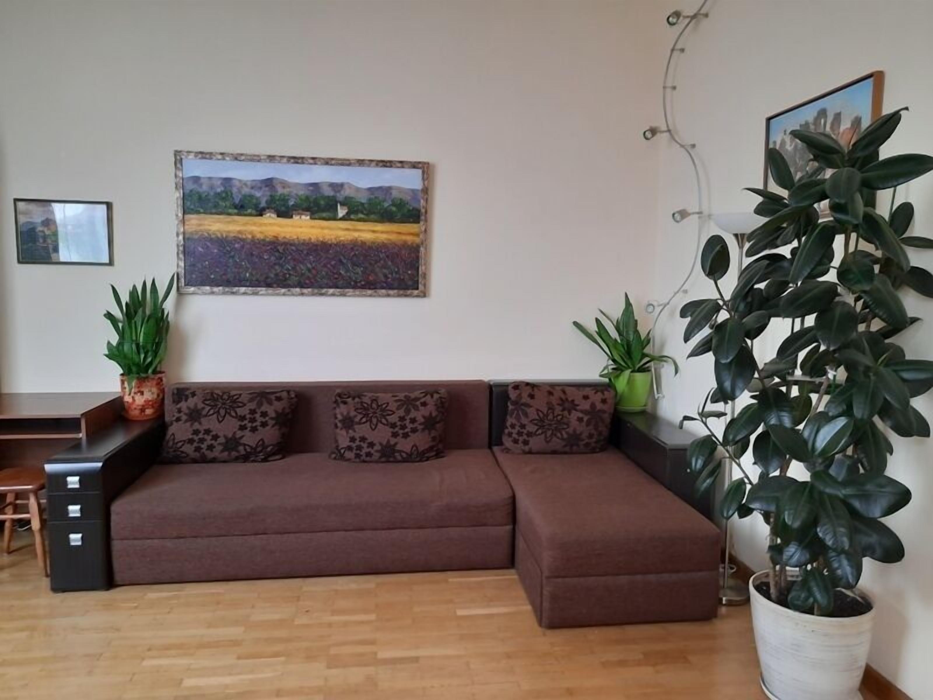 Ein Wohnzimmer mit einem braunen Sofa, zwei Blumentöpfen und einem Bild an der Wand.