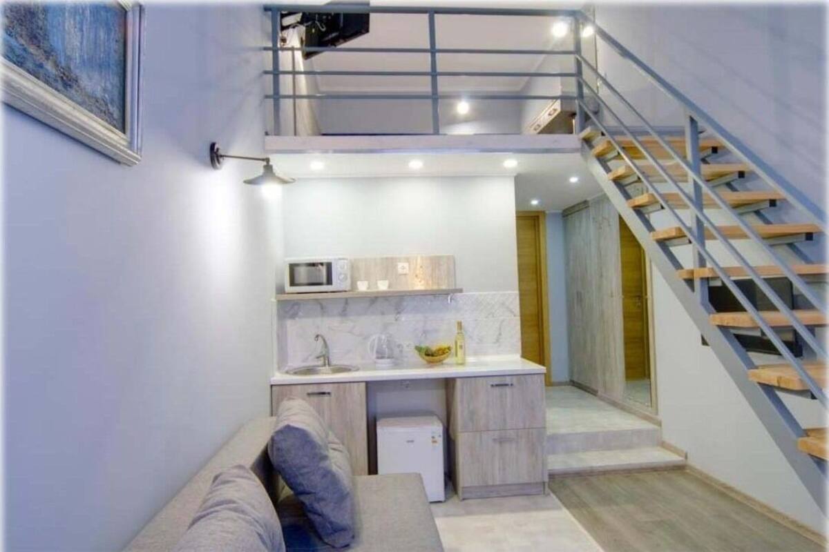 A compact kitchen with a microwave, sink, and cabinets, a small refrigerator, and a wooden staircase.