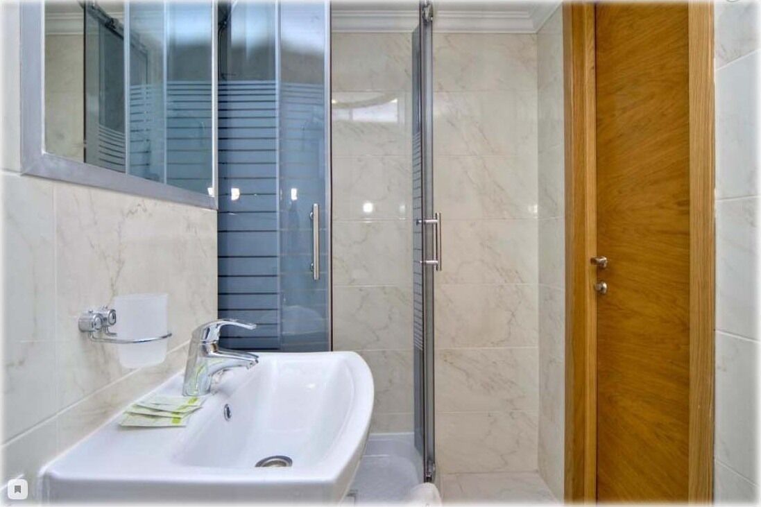 Un baño con lavamanos blanco, espejo, cabina de ducha de vidrio y puerta de madera.