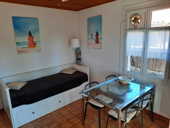 Il s’agit d’une pièce de taille modeste, comprenant un lit, une table à manger en verre et des chaises. Deux tableaux ornent les murs, il y a une fenêtre avec des rideaux et un ventilateur de plafond.