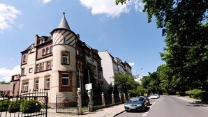 Un edificio histórico con una torre distintiva y esquinas redondeadas, situado en una calle arbolada.