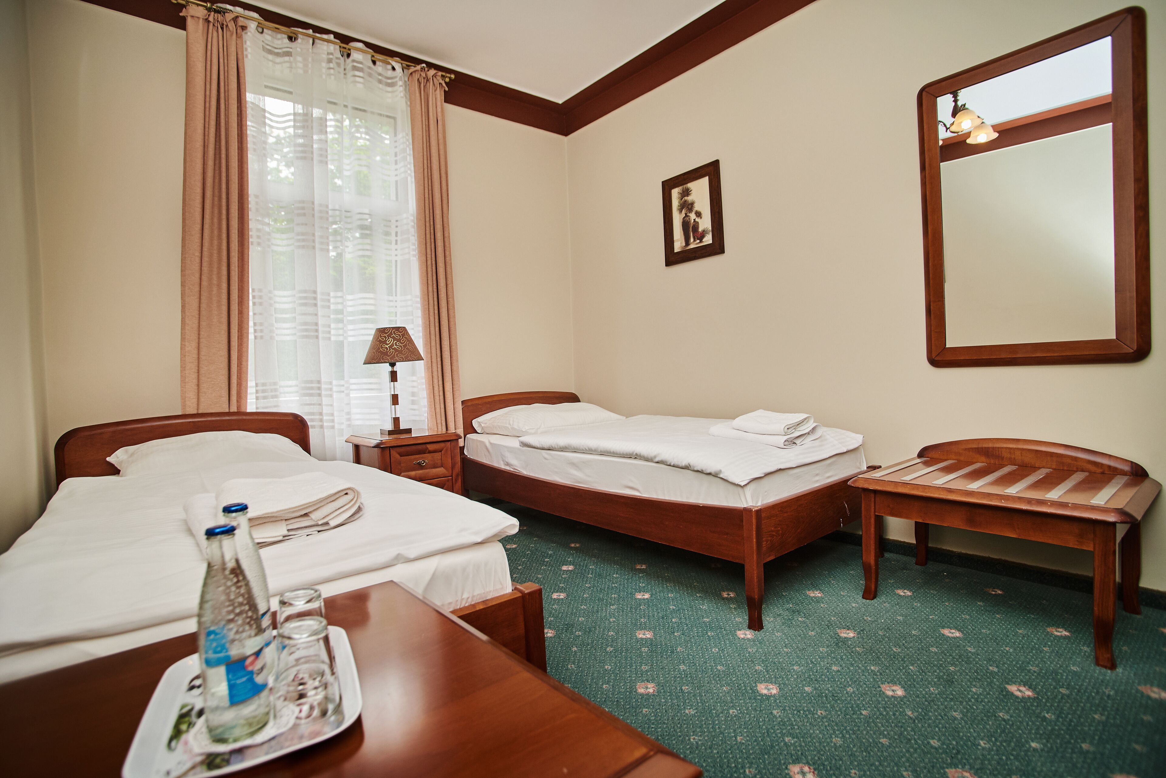 A hotel room with two single beds, a wooden bench, a table with a bottle and glasses, a lamp, and a framed picture on the wall.