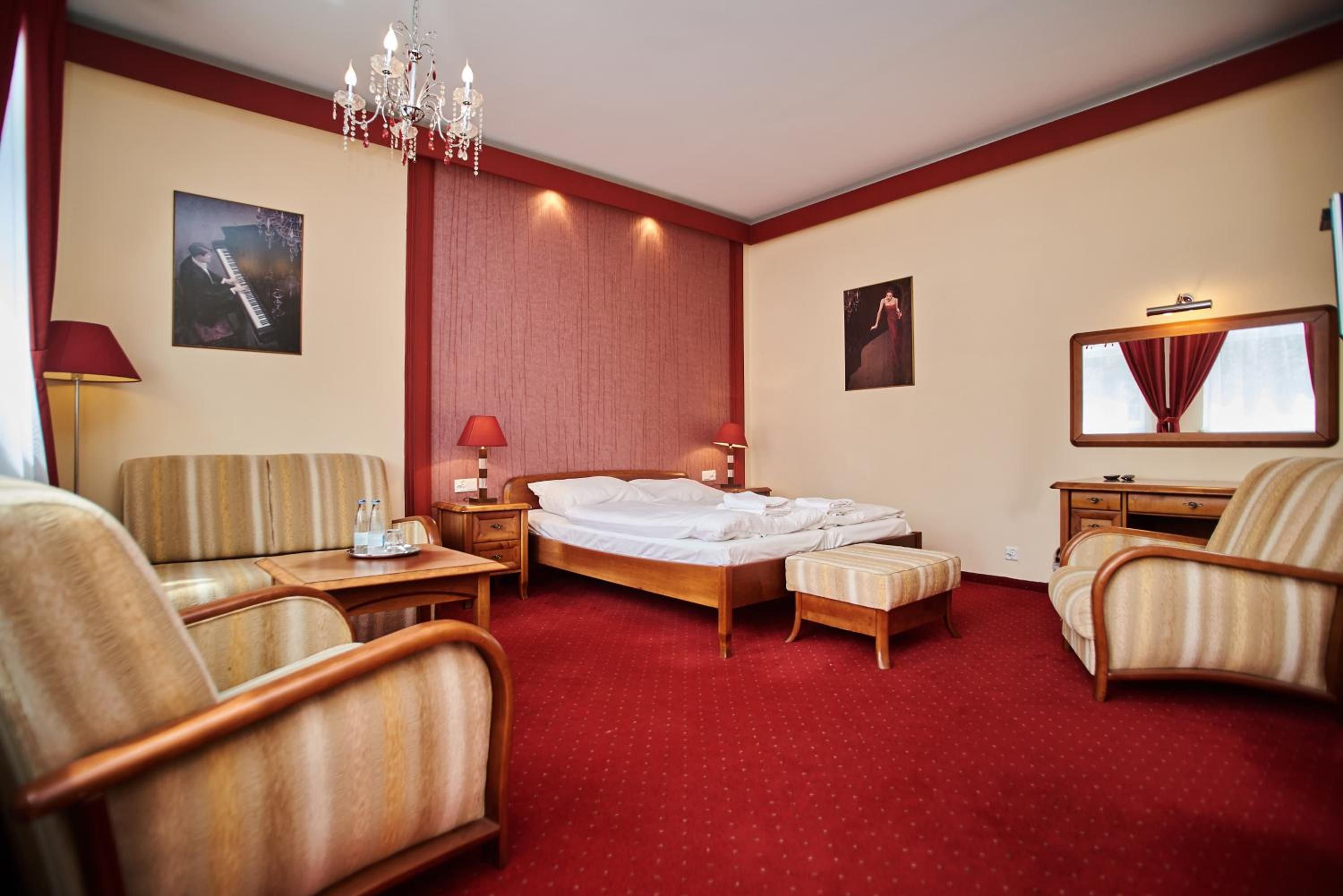A hotel room with a bed, two armchairs, a small table, a desk, a mirror, and a chandelier.