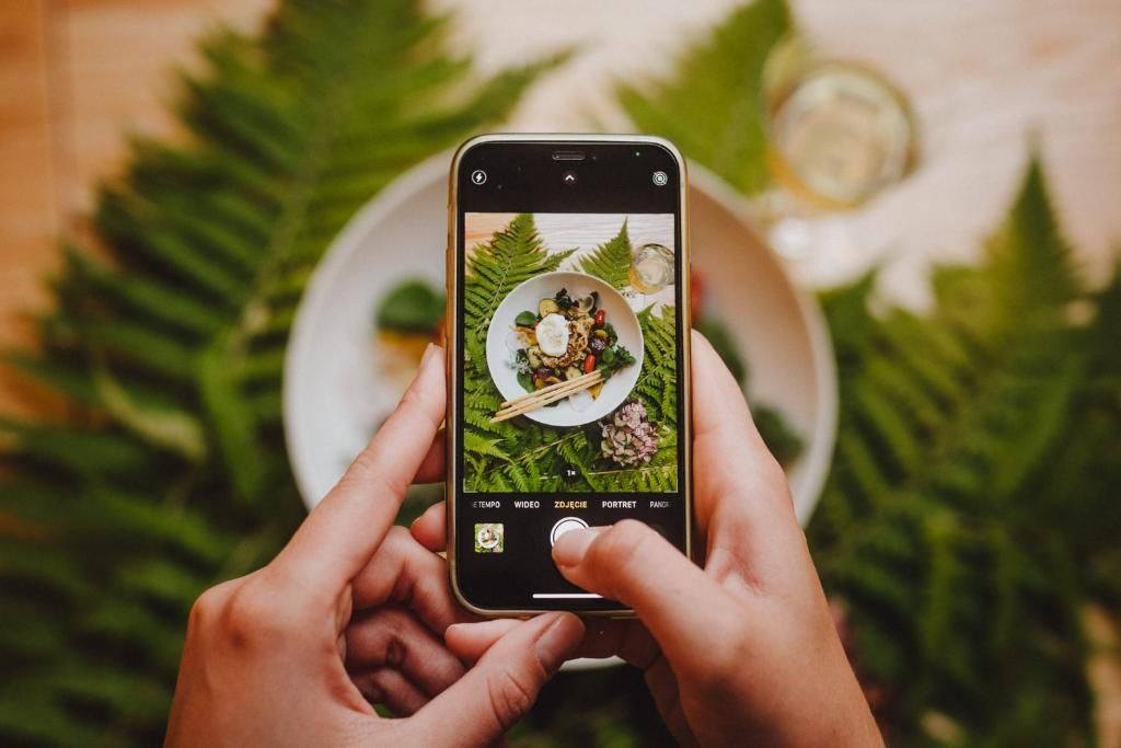 A person holding a smartphone with a photo of a dish on a plate, surrounded by greenery.