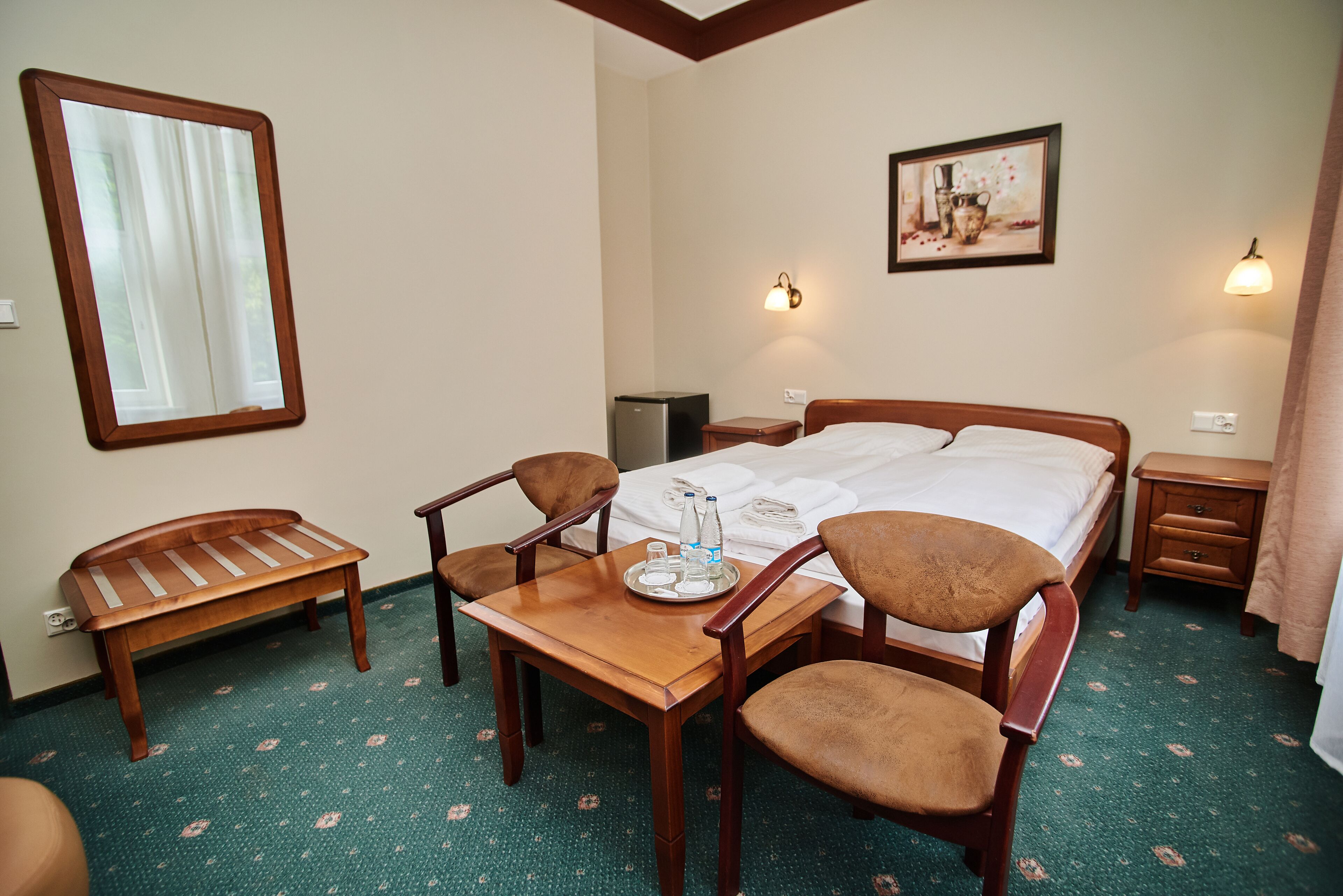 A hotel room with a bed, a wooden table with chairs, a bench, a bedside table with a lamp, and a wall-mounted picture.