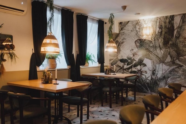 Un comedor con mesas y sillas, un mural en la pared, lámparas colgantes y plantas en macetas.