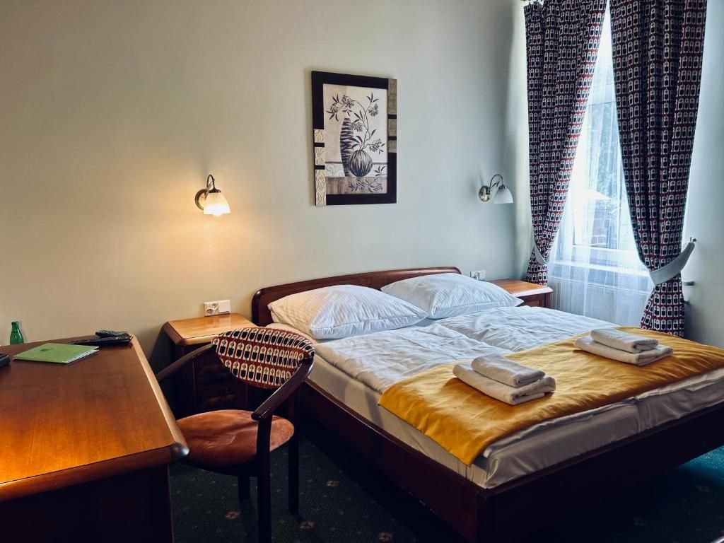 A hotel room with a bed, a desk, a chair, a wall-mounted light, and a window with curtains.