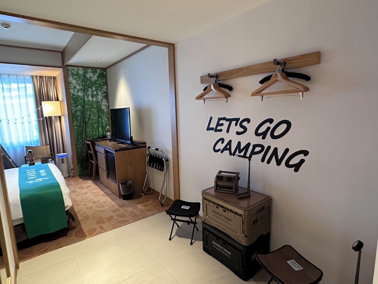 Hotelkamer met een bed, bureau, televisie en een muur met de tekst 'LET'S GO CAMPING'.