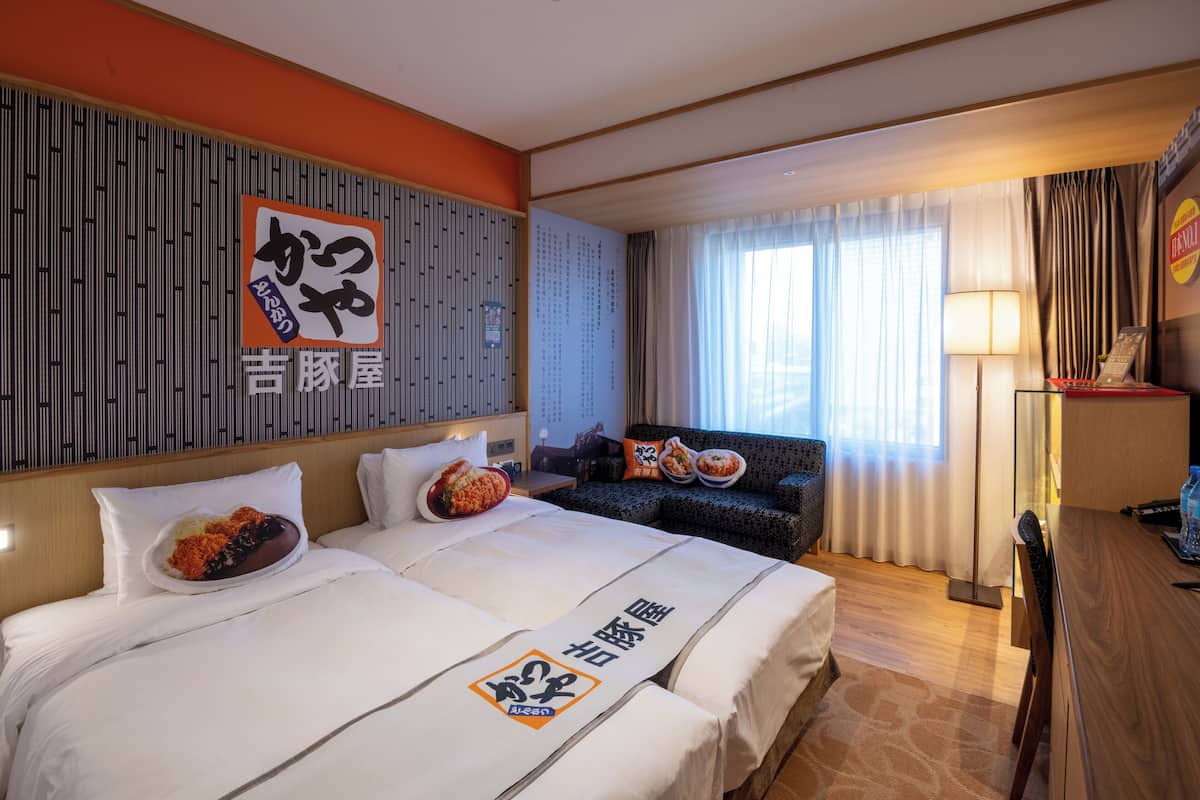 A hotel room with two beds, a sofa, a desk, and a large window with curtains.