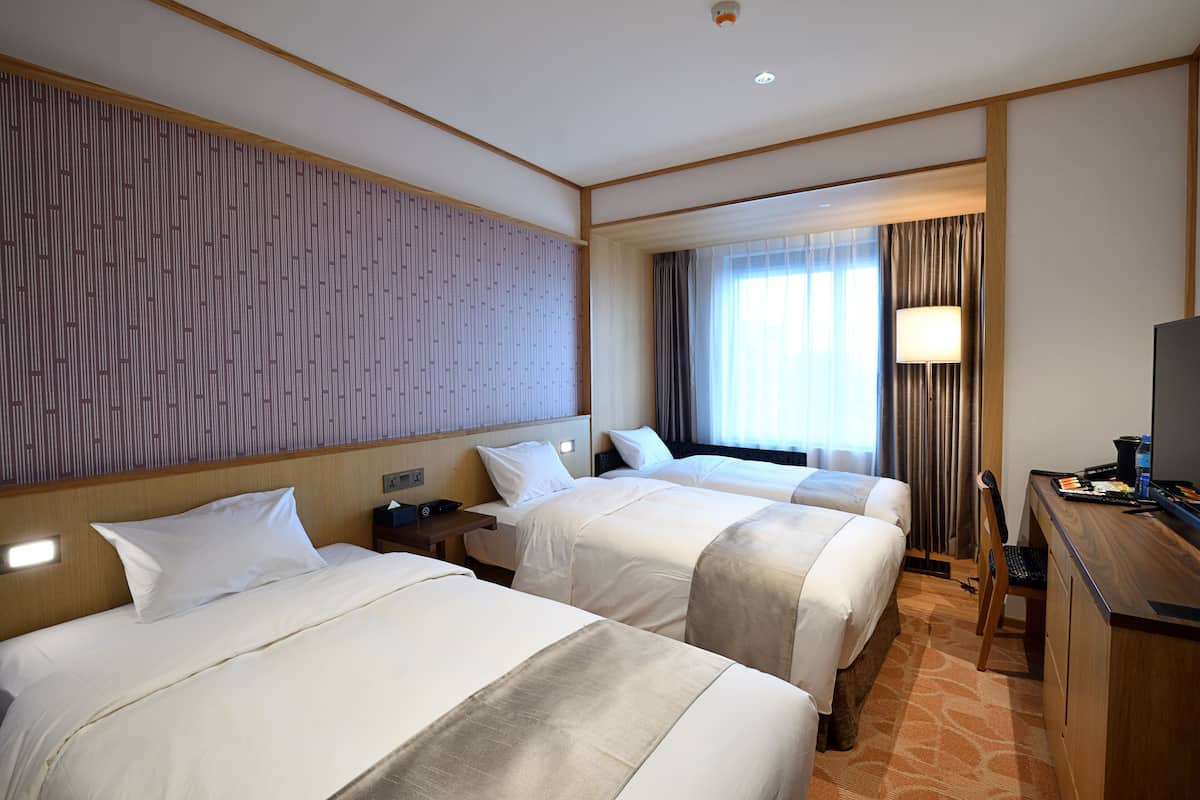 A hotel room with two beds, a desk, a lamp, and a television.