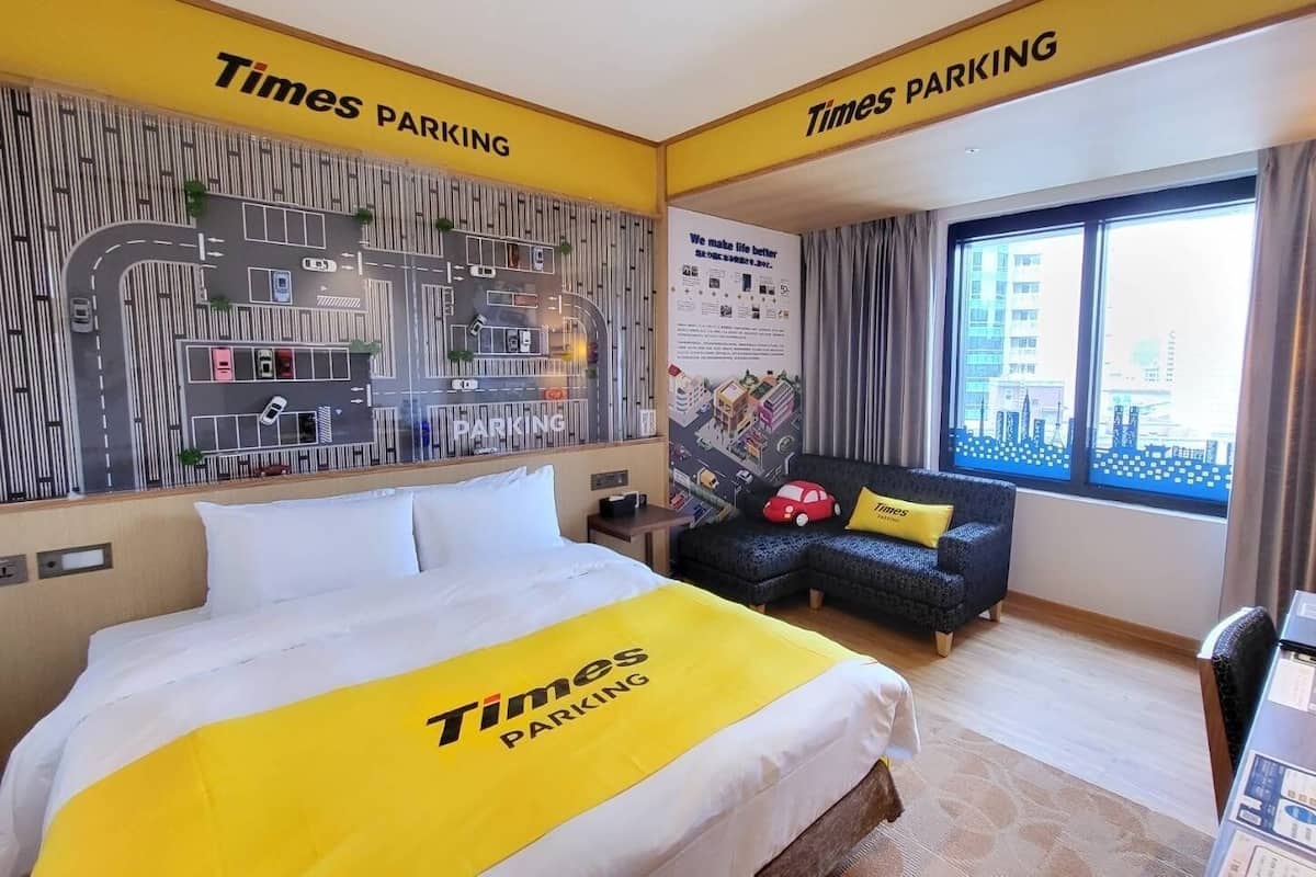 A hotel room with a bed, a desk, a chair, a sofa, a window with a city view, and a Times Parking sign.
