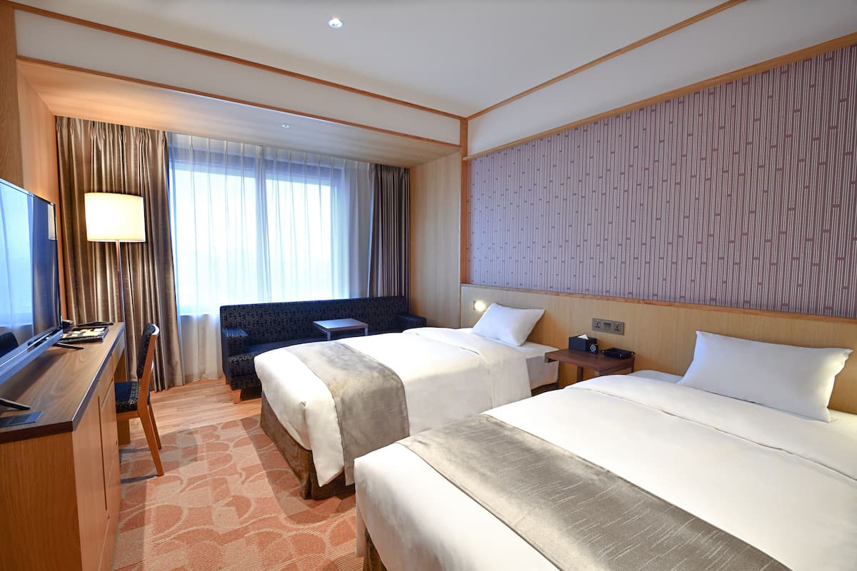 A hotel room with two beds, a desk, a chair, a TV, and a large window with curtains.