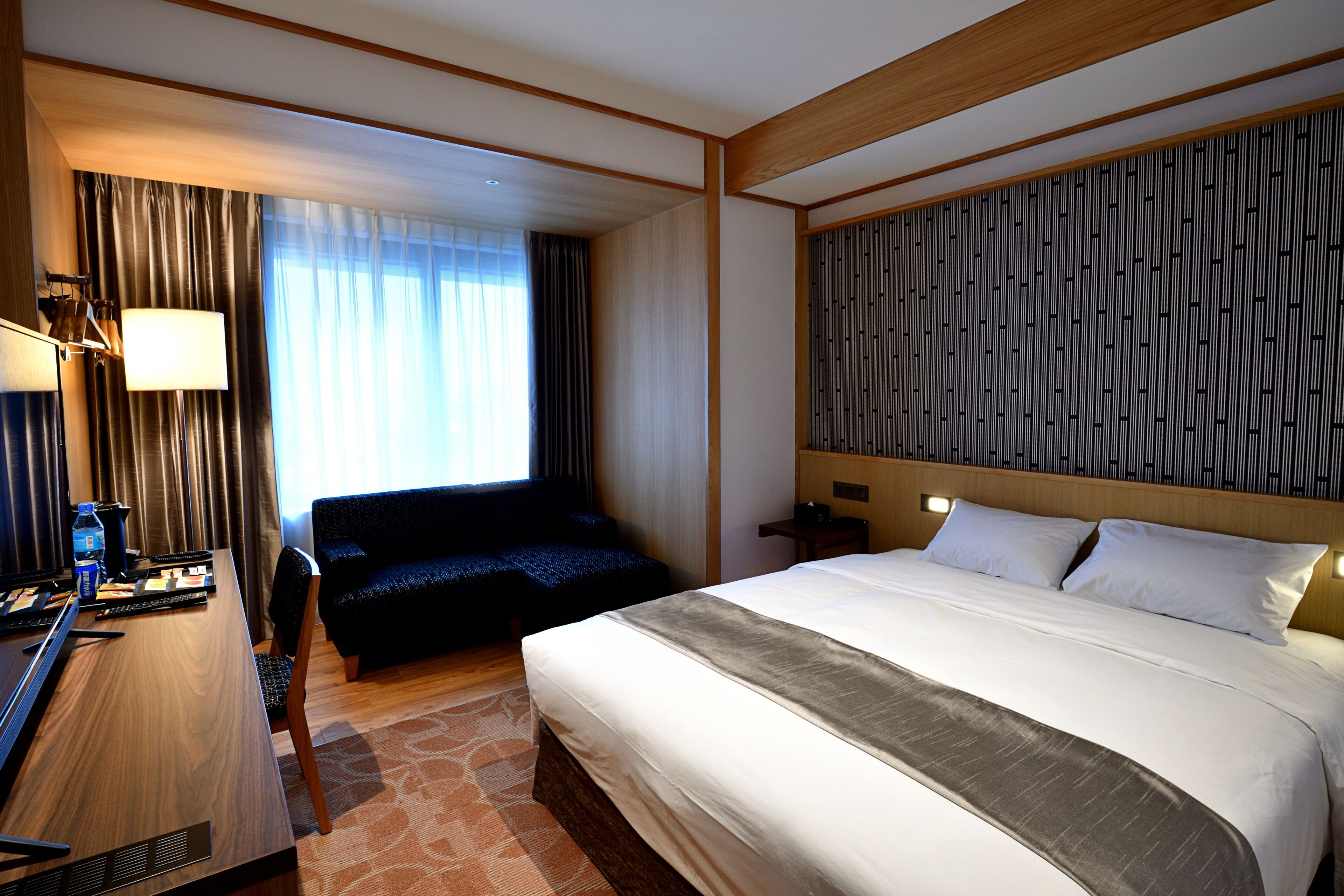 A hotel room with a large bed, a desk with a computer, a chair, a sofa, and a patterned headboard.