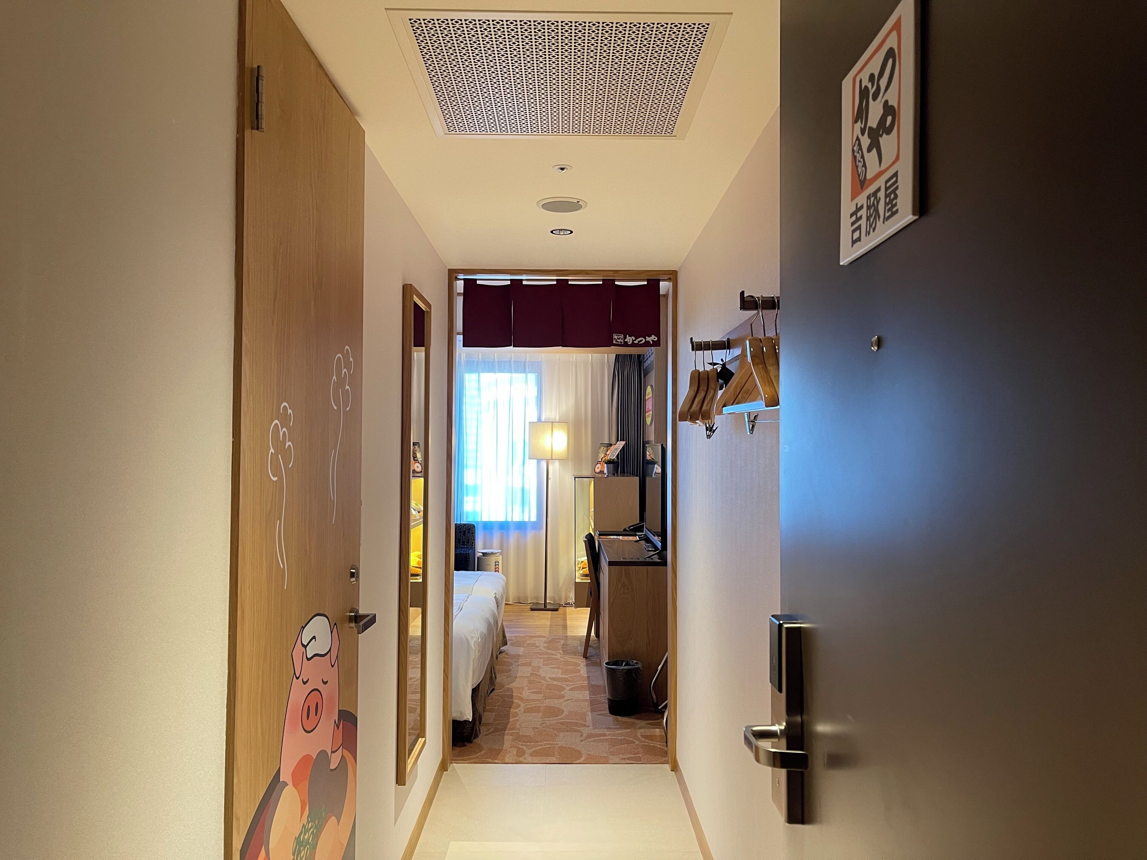 A hotel corridor with a wooden door with a pig mural, a bed, a desk, and a shoe rack.
