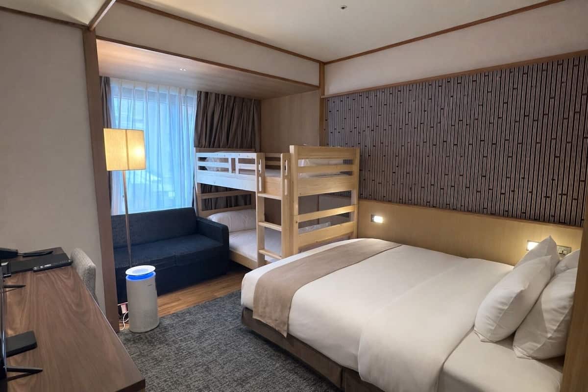 A hotel room with a bunk bed, a desk with a chair, a lamp, and a view of the outdoors.