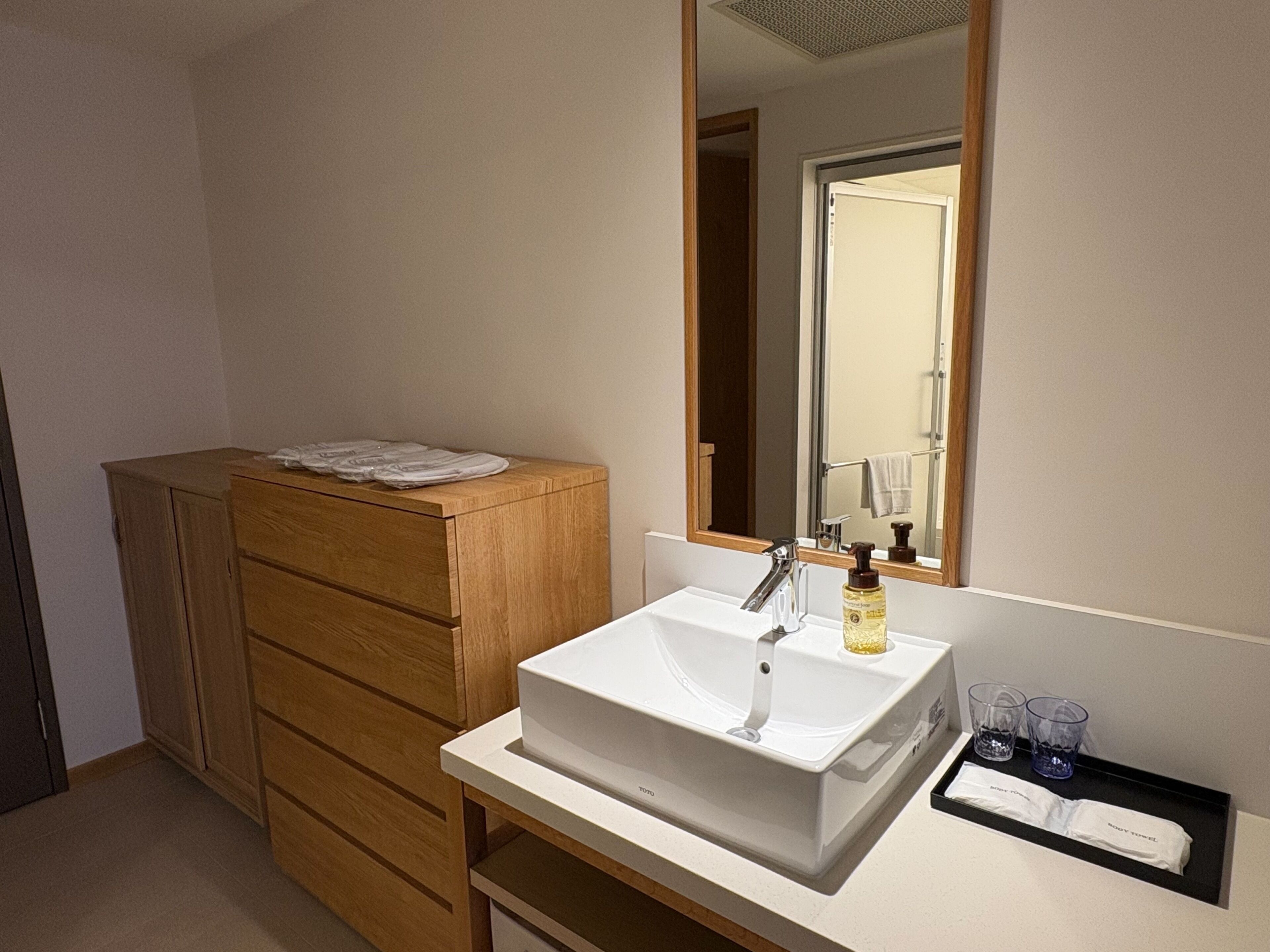 A bathroom with a wooden cabinet, a white sink, a mirror, and a tray with a towel and glasses.